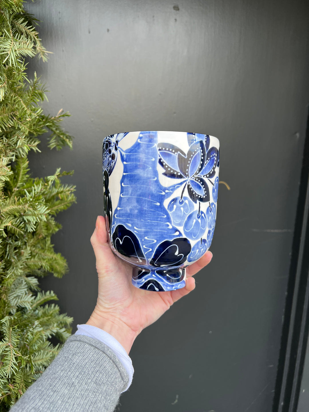 A large wide vase with a white background and blue hand painted pattern of fruit bearing branched on it. It has a gold rim.