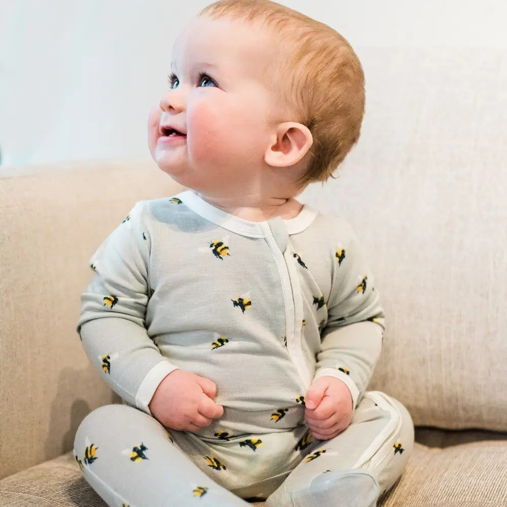 A baby sits smiling while wearing a light blue zipped romper with a bee print. 