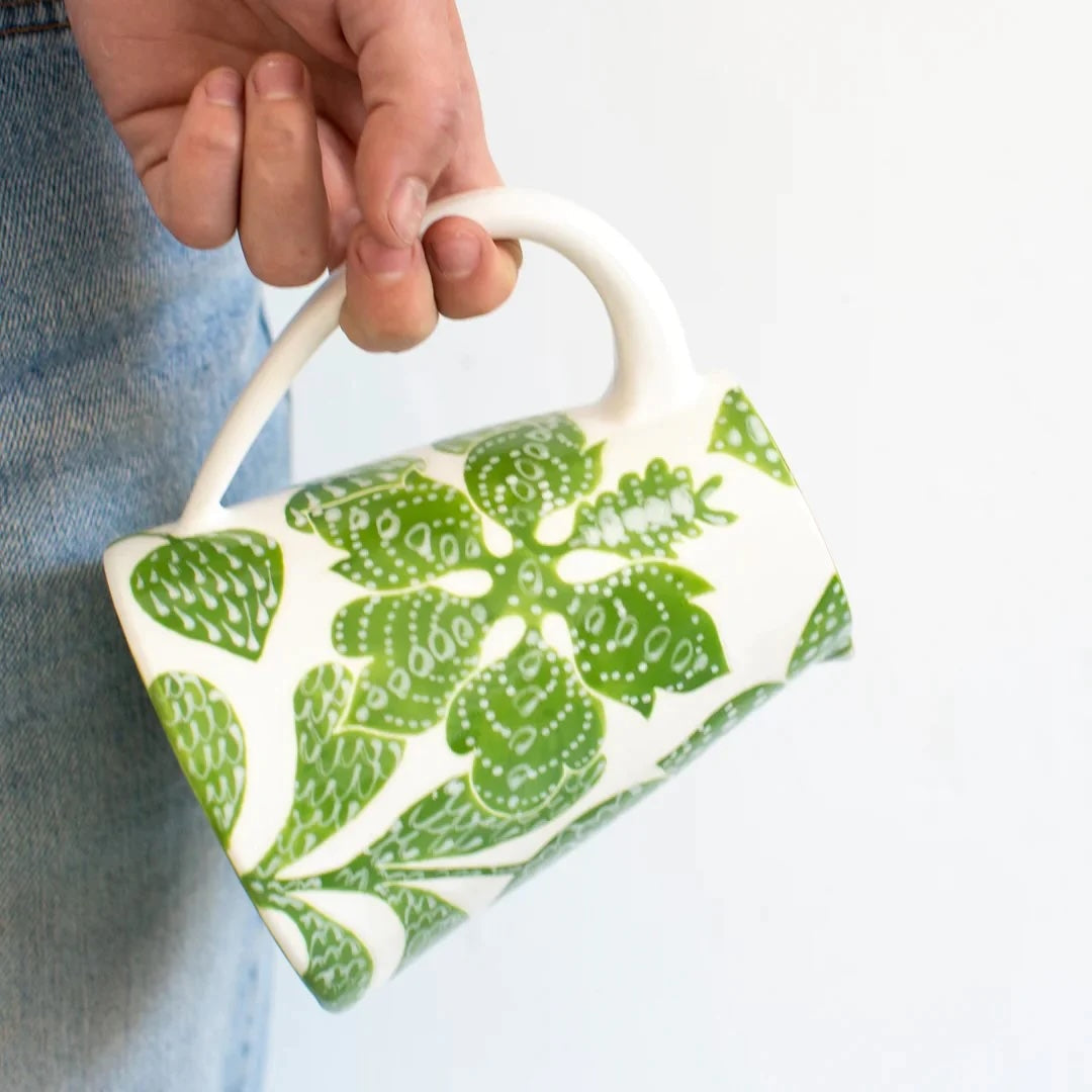 A cream colored small pitcher with a green leaf design hangs from someone's hand.