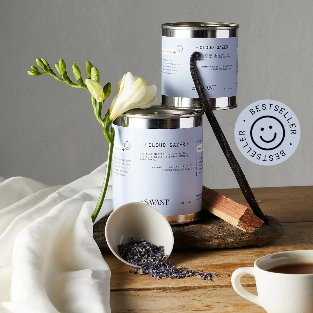 Two candles in aluminum tins with light blue sleeves surrounded by lavender, a cup of tea and a lily.