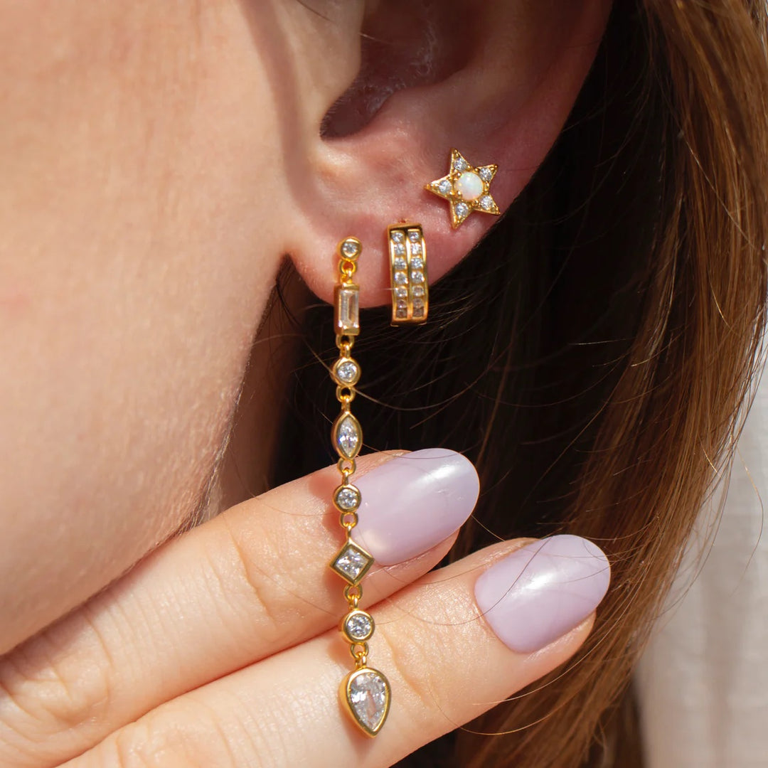 A woman points out long gold colored straight earrings with diamond like gems on it. She also wears a small hoop and stud.