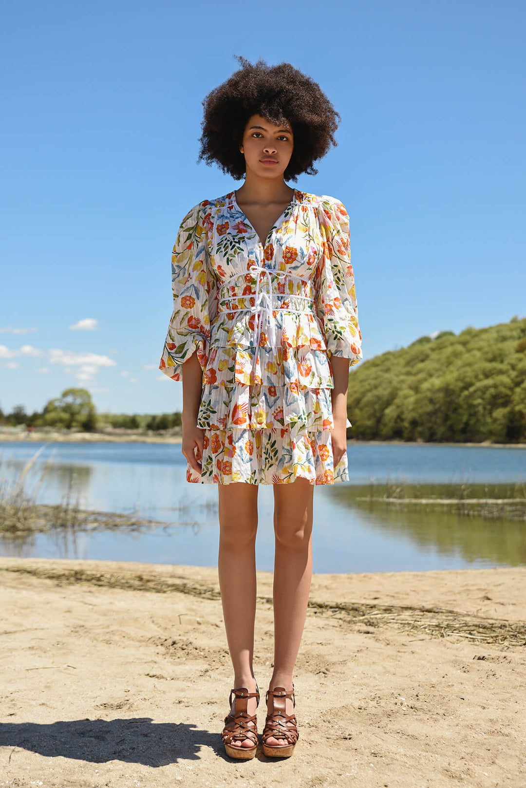 A lady with long legs stands by a lake while wearing a 3/4 sleeve mini dress with a floral print.