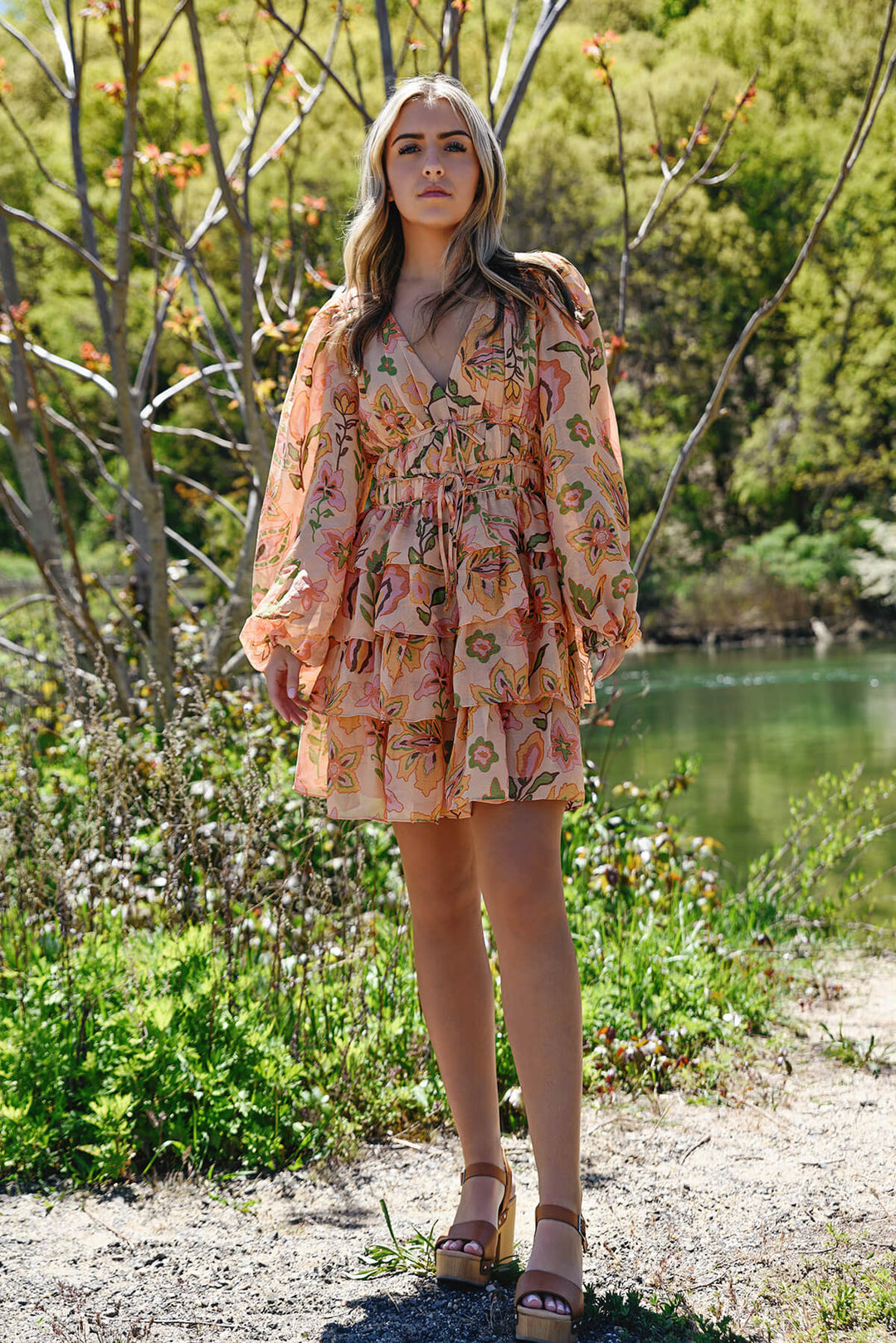 A lady stands next to a lake in a long sleeved min dress with a tiered skirt and floral print.