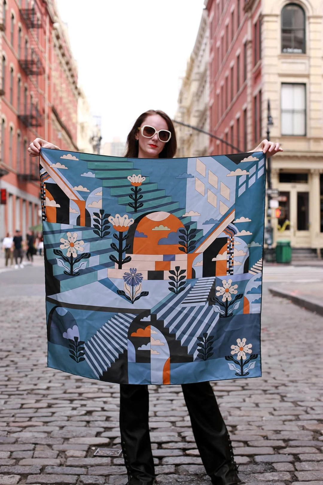A lady standing on a cobblestone street in Soho holds a large multi pattern, multi color scarf in front of her.