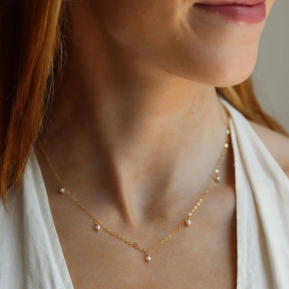 A woman wears a gold necklace with 5 small delicate freshwater pearls attached.